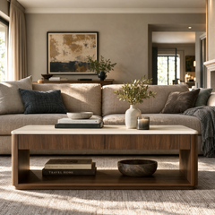 A living room with a beige sofa behind a low rectangular coffee table, topped with books, a vase of greenery, a bowl, and a candle.