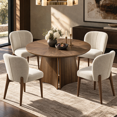 A round wooden dining table surrounded by four cream upholstered chairs, set with flowers, candles, and a bowl of figs in a sunlit room.