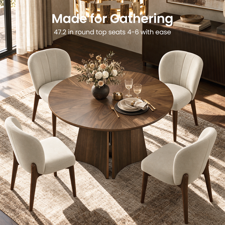 Round brown dining table with a radial grain pattern and broad pedestal base, surrounded by four cream chairs and set with flowers, two glasses and a place setting.