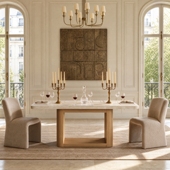 A dining table set with candlesticks, wine glasses, and a decanter sits between two upholstered chairs beneath a chandelier and wall artwork, with tall arched windows on either side.