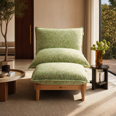 Lounge chair with three oversized pale green textured cushions and a light brown frame, flanked by tables beside a large window in a sunlit room.