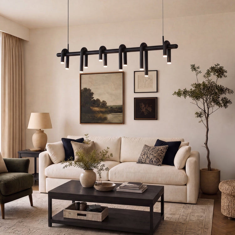 A black pendant light with curved tubular arms hangs above a cream sofa in a living room with framed artwork, a dark coffee table, a table lamp and a tall potted tree.