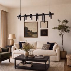 Black linear pendant light with curved tubular arms and glowing ends hangs above a cream sofa in a living room with framed artwork, a dark coffee table and a tall potted tree.