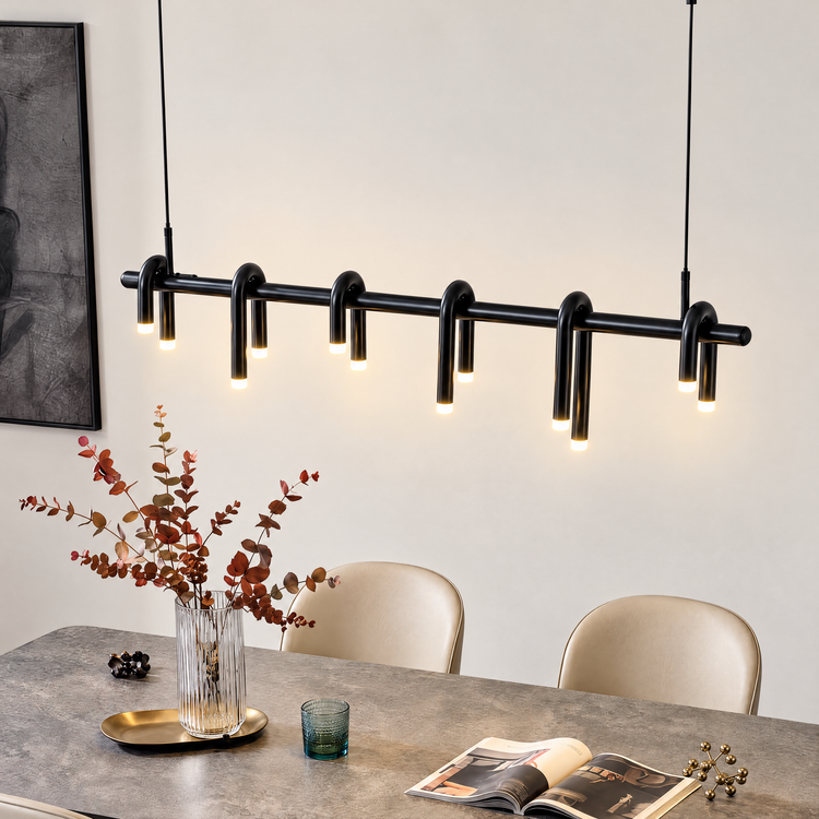 Black pendant light with six curved, double-ended illuminated tubes along a horizontal bar, suspended above a dining table with a vase of reddish branches and cream chairs.