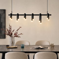 Black pendant light with six curved tubular loops and glowing tips hangs above a gray dining table with cream chairs, a vase of branches, a blue drinking glass and an open magazine.