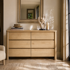 A light wood-grain six-drawer dresser sits below a framed mirror with books, sculpture, and candle