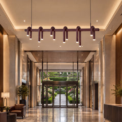 A burgundy pendant light with looping tubes and glowing downward-facing ends hangs in a tall lobby with glossy floors, seating and glass doors overlooking a garden.
