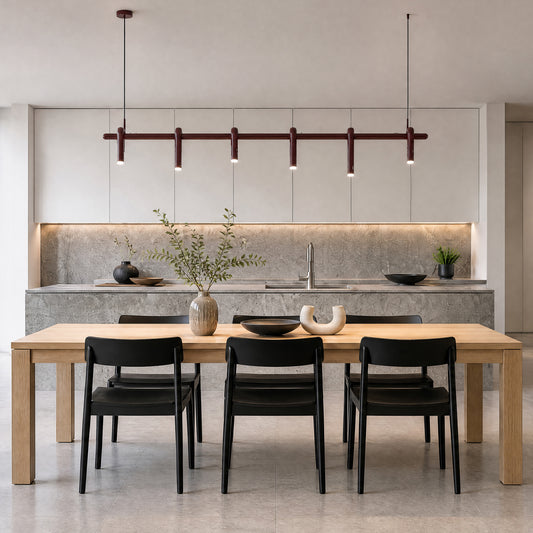 A dark red linear pendant with six downward-facing lights hangs above a light brown dining table with black chairs in a kitchen with pale cabinets and gray counters.