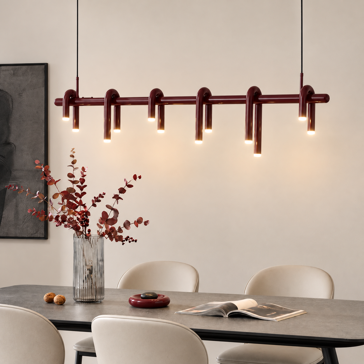 Glossy burgundy pendant light with curved tubular arms and glowing downward-facing ends hangs above a gray dining table with cream chairs, a vase of reddish foliage and an open book.