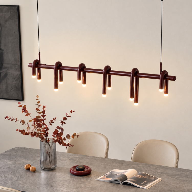 A burgundy pendant light with six inverted U-shaped tubes and glowing tips hangs above a gray dining table with a vase of reddish foliage and an open magazine.