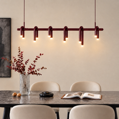 Burgundy pendant light with six curved loops and glowing tips hangs above a dining table with cream chairs, a vase of reddish foliage, and an open book.