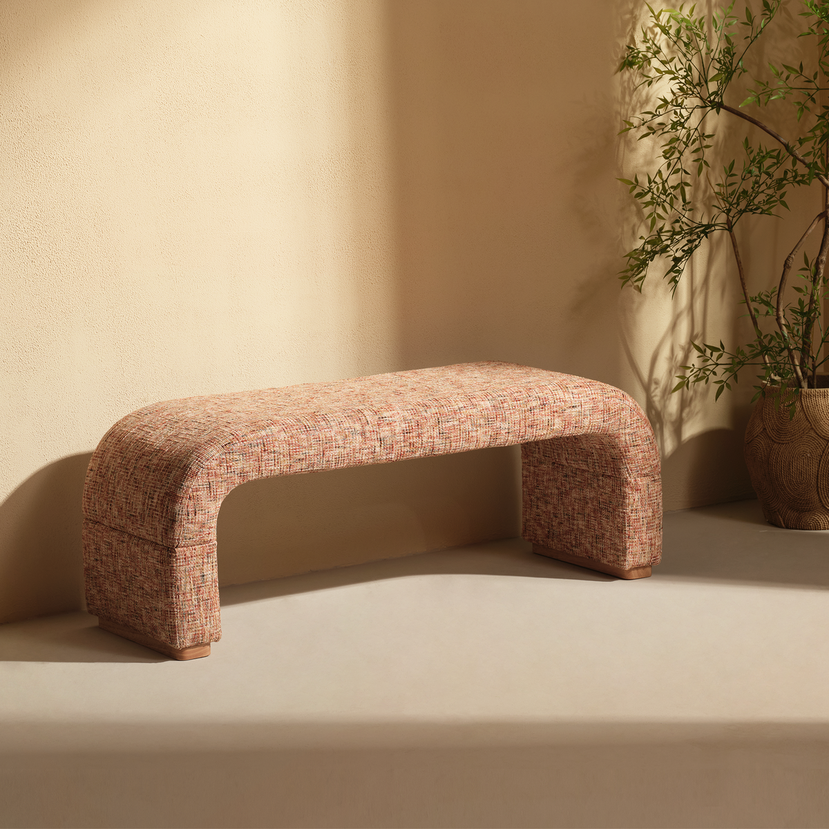 Reddish textured upholstered bench with a curved arch-shaped profile and short wood-tone feet beside a plant