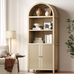 Light wood-tone arched bookcase with open display shelves and fluted lower doors beside a living room chair