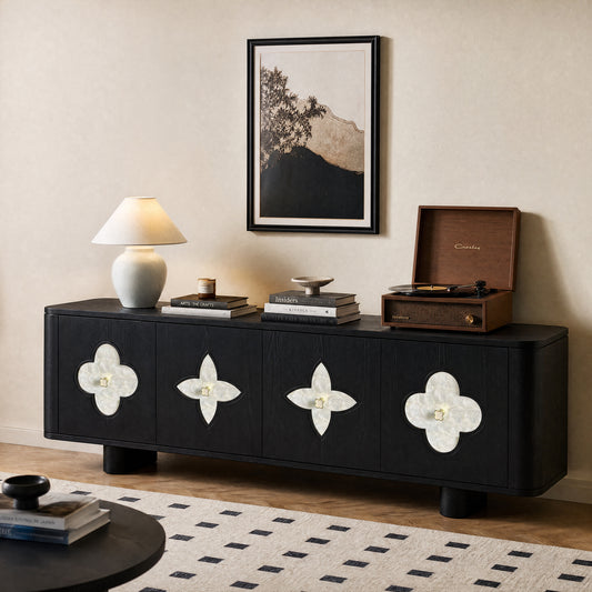 A black sideboard with four pale clover-shaped door insets stands beneath framed landscape art; a lamp, books, bowl, and record player sit on top.