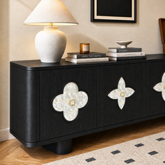 Black sideboard with rounded corners, pale four-lobed door insets and gold-colored knobs, topped with a lit cream lamp, stacked books, an amber candle jar and a small pedestal bowl.