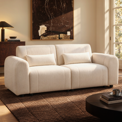 A cream two-seat sofa with two cushions sits on a dark textured rug in a sunlit living room, beneath a large abstract painting.