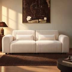 Cream sofa with rounded arms and two rectangular cushions sits on a brown rug beneath framed abstract artwork, with a table lamp to the left and sunlight across the wall.