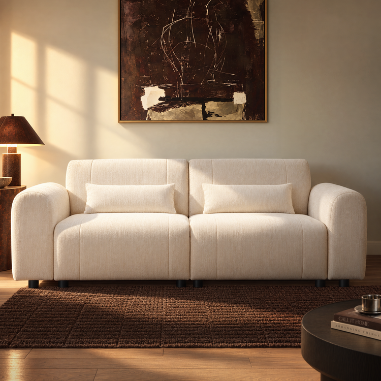 Cream sofa with rounded arms and two rectangular cushions sits on a brown rug beneath framed abstract artwork, with a table lamp to the left and sunlight across the wall.