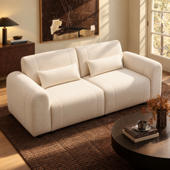A cream-colored sofa with two matching cushions sits on a dark textured rug in a sunlit living room.