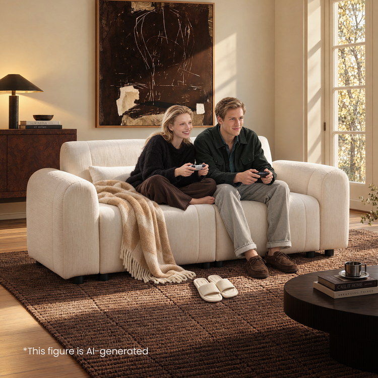 Two people holding game controllers sit on a cream sofa with rounded arms and a draped blanket in a sunlit room with abstract wall art and a brown rug.