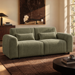 Olive green sofa with two matching cushions in a sunlit living room, with abstract wall art, a rug, and a coffee table.