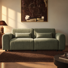 Muted green sofa with rounded arms, two seat cushions and two rectangular pillows, set on a brown rug beneath an abstract painting beside a lit table lamp.