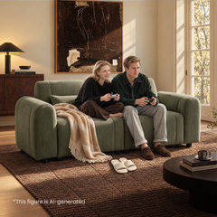 Two people hold game controllers while sitting on a green sofa with rounded arms and a beige throw in a sunlit living room with abstract wall art and a brown rug.