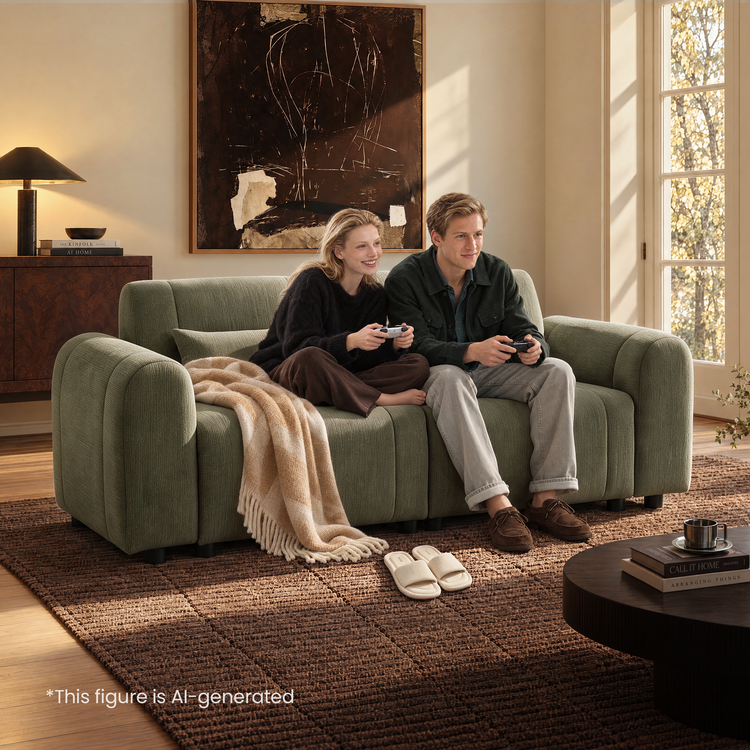 Two people hold game controllers while sitting on a green sofa with rounded arms and a beige throw in a sunlit living room with abstract wall art and a brown rug.