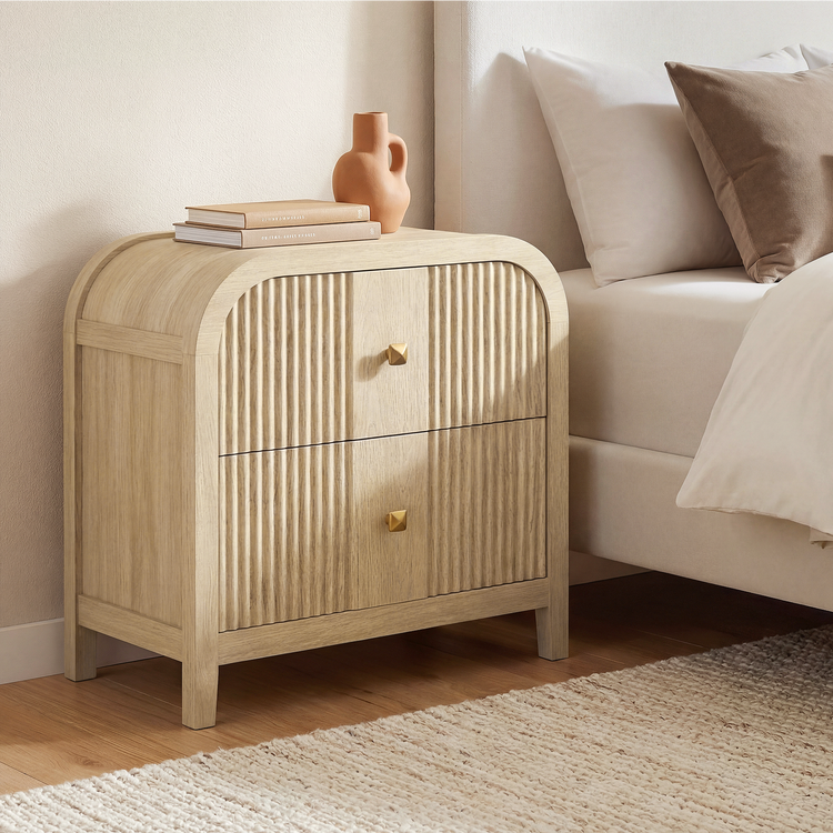 A light wood bedside cabinet with a rounded arched top, two fluted drawers, and brass knobs stands beside a bed