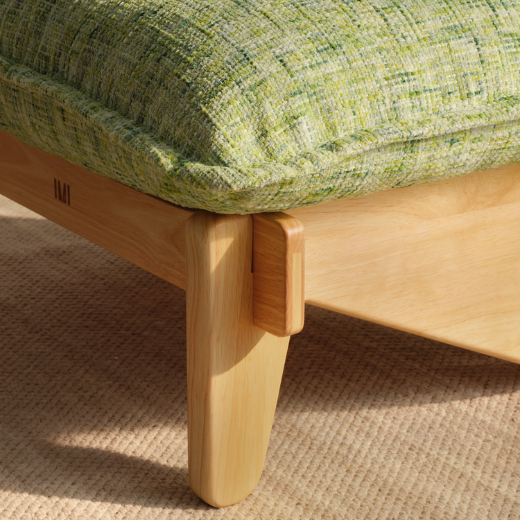 Close-up of a green and cream textured cushion with piped edges on a light wood frame with a rounded leg, standing on a beige rug.