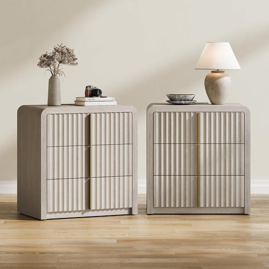 Two light-colored chests with three vertically fluted drawer fronts stand side by side, topped with a vase, books, a camera, bowls, and a lamp.