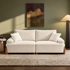 Cream sofa with two seat cushions, rounded arms and two throw pillows on a textured rug, beneath framed artwork on an olive-green wall, with a side table and floor lamp.