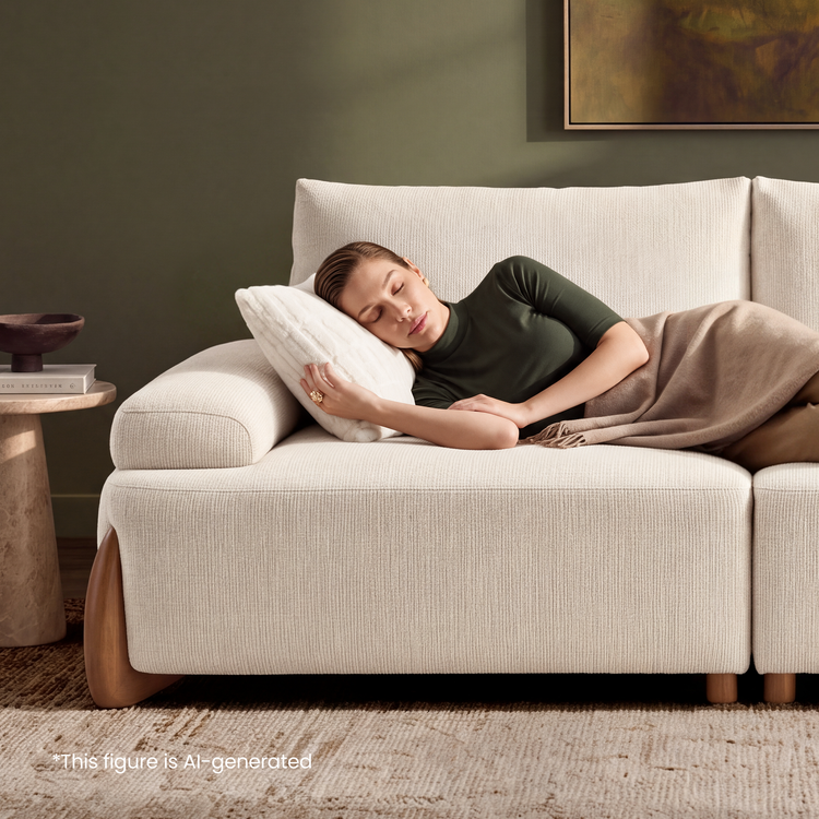 A woman rests on her side on a cream sofa, hugging a white pillow and covered by a taupe blanket.