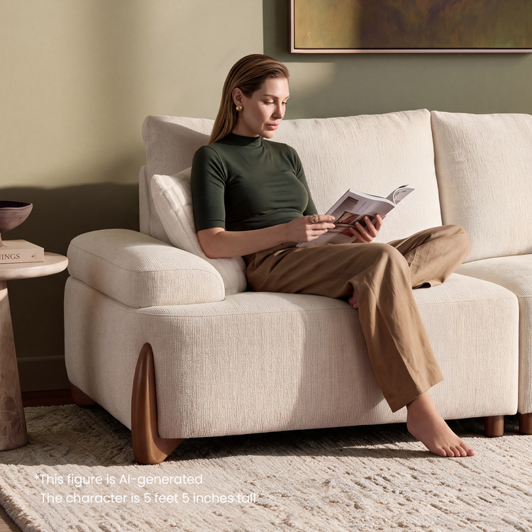 A woman sits barefoot on a cream sofa reading an open magazine, with a rug, side table, and framed artwork in the room.