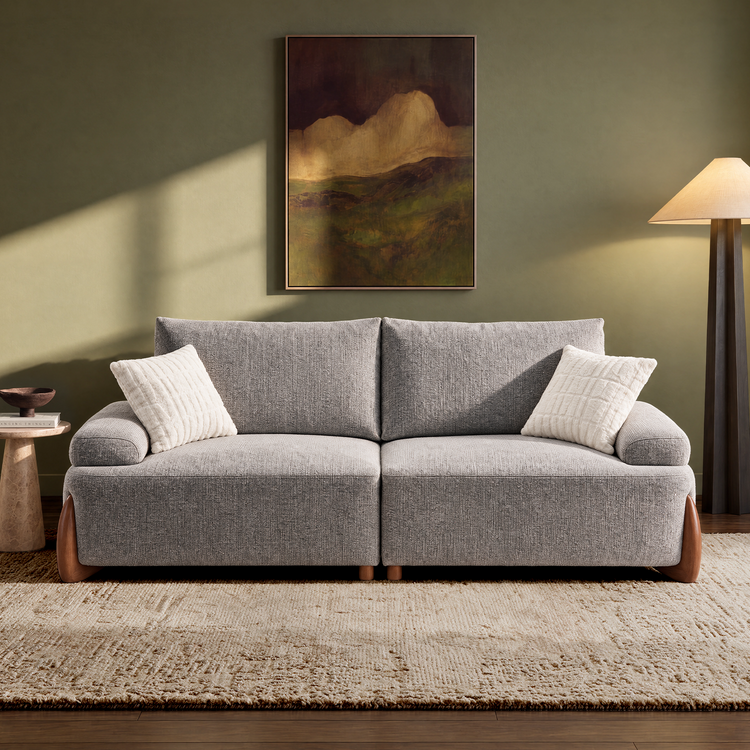 A gray two-seat sofa with two white pillows sits on a cream rug beneath a framed landscape painting; a floor lamp stands to the right.