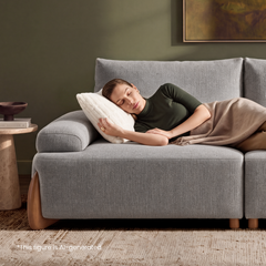 A woman with closed eyes rests on a gray sofa, her head on a white pillow and a beige blanket over her legs, beside a small table holding books and a bowl.