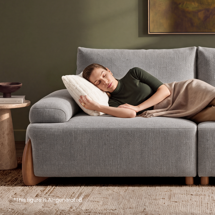 A woman with closed eyes rests on a gray sofa, her head on a white pillow and a beige blanket over her legs, beside a small table holding books and a bowl.