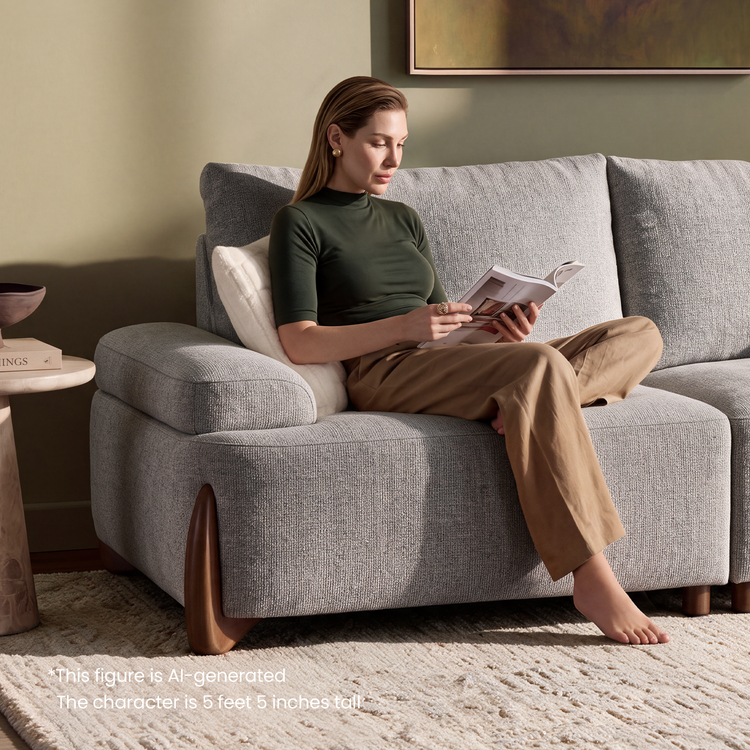 A woman sits cross-legged on a gray sofa, reading a magazine in a living room.