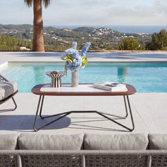A low rounded coffee table with a brown top and black bent-wire legs stands beside a swimming pool