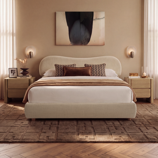 A neatly made bed with a rounded upholstered headboard, patterned and solid dark pillows, and a brown blanket, flanked by wooden nightstands and wall lights.
