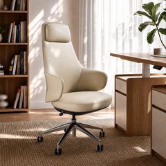 Cream-colored office chair with a tall back, padded headrest, curved armrests and wheeled base beside a desk in a sunlit room with bookshelves and a leafy plant.