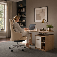 A woman sits in a cream high-backed office chair using a laptop at a desk with drawers, open shelves, a gold-colored lamp, a mug and a potted plant.