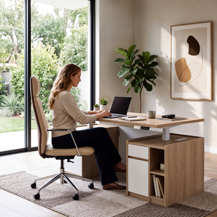 A woman sits in a cream office chair using a laptop at a desk with drawers and open shelves, beside tall windows, green plants and framed abstract art.