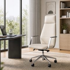 Cream high-back office chair with silver-colored arm supports and a five-spoke wheeled base beside a dark desk, with large windows and shelving in the background.