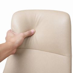 A hand presses a thumb into a beige padded chair back, creating an indentation beside its rounded edge and curved horizontal seam.