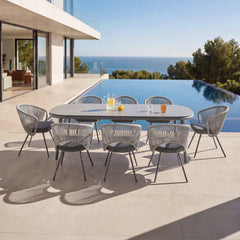 Eight gray rope-back chairs surround a long outdoor dining table, The patio overlooks a pool and ocean