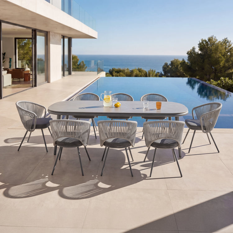 Eight gray rope-back chairs surround a long outdoor dining table, The patio overlooks a pool and ocean