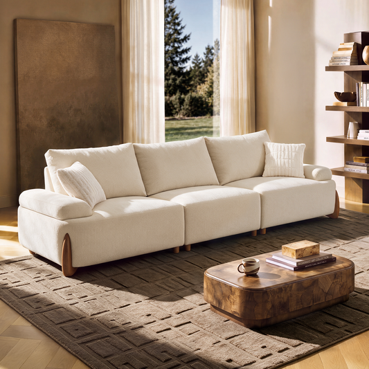 A cream-colored sofa with two cushions sits on a textured rug beside a wooden coffee table in a sunlit living room.