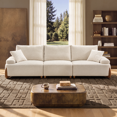 A cream three-seat sofa with two pillows sits in a sunlit living room, with a coffee table, patterned rug, bookshelves, and a window overlooking trees.