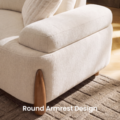 Close-up of a cream-colored sofa with a broad rounded armrest, textured upholstery, a brown tapered leg, and a fluffy cushion on a patterned rug.